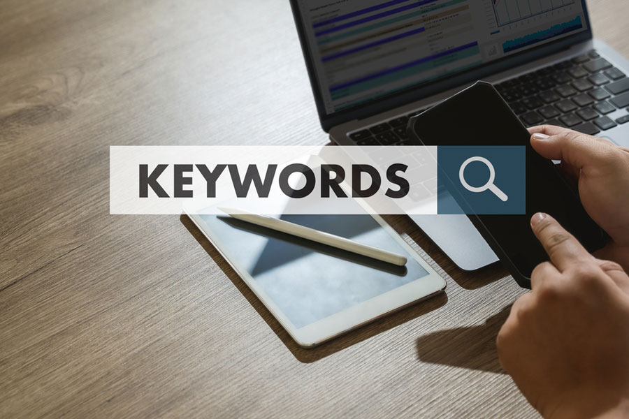 Word keywords on search bar and hands on cell with tablet and laptop computers on desk.