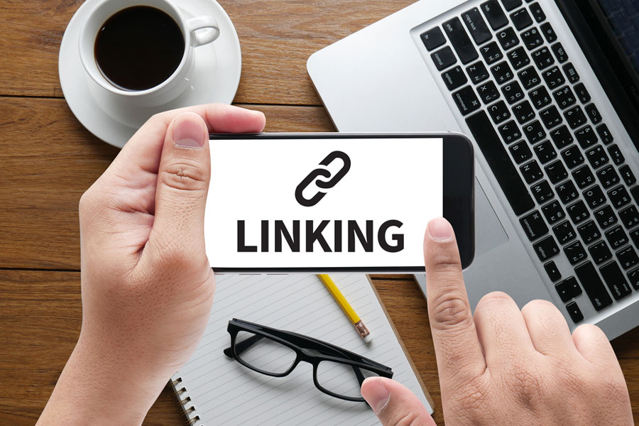 Two hands holding mobile phone with word linking on it and laptop in background.